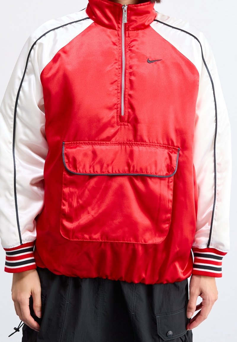 Person klädd i en röd och vit sportig zip-up-jacka med framficka, svarta byxor och randiga muddar på ärmarna.