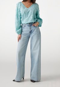 Blouse bleu clair, texturée avec des détails floraux et des liens au niveau du col, associée à un jean en denim clair à taille haute et à jambes larges, et des talons noirs.