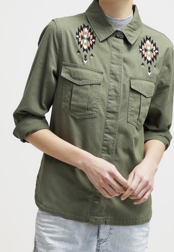 Camisa de color verde oliva, de botones y con mangas cortas, que cuenta con dos bolsillos frontales y patrones geométricos bordados en rosa y negro en el pecho.
