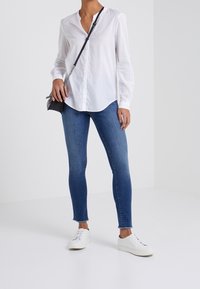 Blusa blanca de manga larga con cuello redondo, combinada con jeans ajustados azules. Modelo lleva zapatillas blancas y un pequeño bolso cruzado negro.