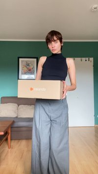 Ruskea Zalando-laatikko, jota pitää henkilö mustassa hihattomassa paidassa ja vaaleanharmaissa leveälahkeisissa housuissa huoneessa, jossa on vihreä seinä.