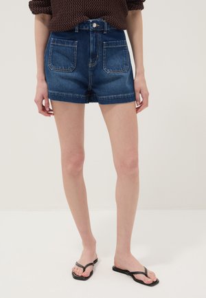 Persoon die donkerblauwe denimshorts draagt met voorzakken, een bruine gebreide trui en zwarte teenslippers, staand tegen een witte achtergrond.