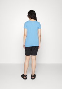 Kvinde står vendt væk iført en lyseblå kortærmet skjorte, sorte opsmøgede shorts og sorte sandaler på en ensfarvet baggrund.