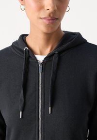 Hanorac zip-up negru, confecționat dintr-un material moale. Are glugă cu șnur, zipper argintiu și manșete texturate. Design simplu, fără modele.