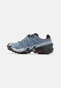 Chaussure de trail running, upper synthétique en bleu clair avec des accents sombres, semelle extérieure en caoutchouc noir, motif de traction unique et système de laçage rapide.