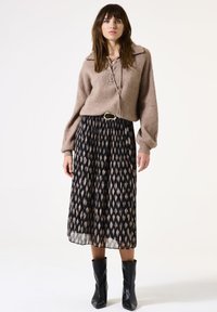 Frau in einem braunen Strickpullover mit Schnürung, schwarzem, gemustertem, plissiertem Midikleid, schwarzem Gürtel und schwarzen Stiefeletten, die vor einem weißen Hintergrund steht.