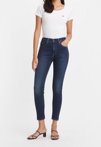 Högmidjade mörkblå jeans med slim fit, tillverkade av denimtyg. Kombinerade med en vit T-shirt och svarta sandaler med remmar.