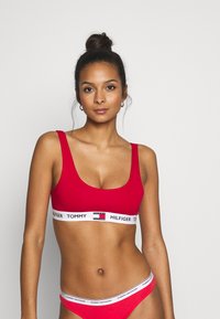 Bralette rossa con una ampia fascia elastica bianca con il testo "TOMMY HILFIGER" in nero. Design semplice con scollatura rotonda e texture liscia. Slip rossi coordinati.