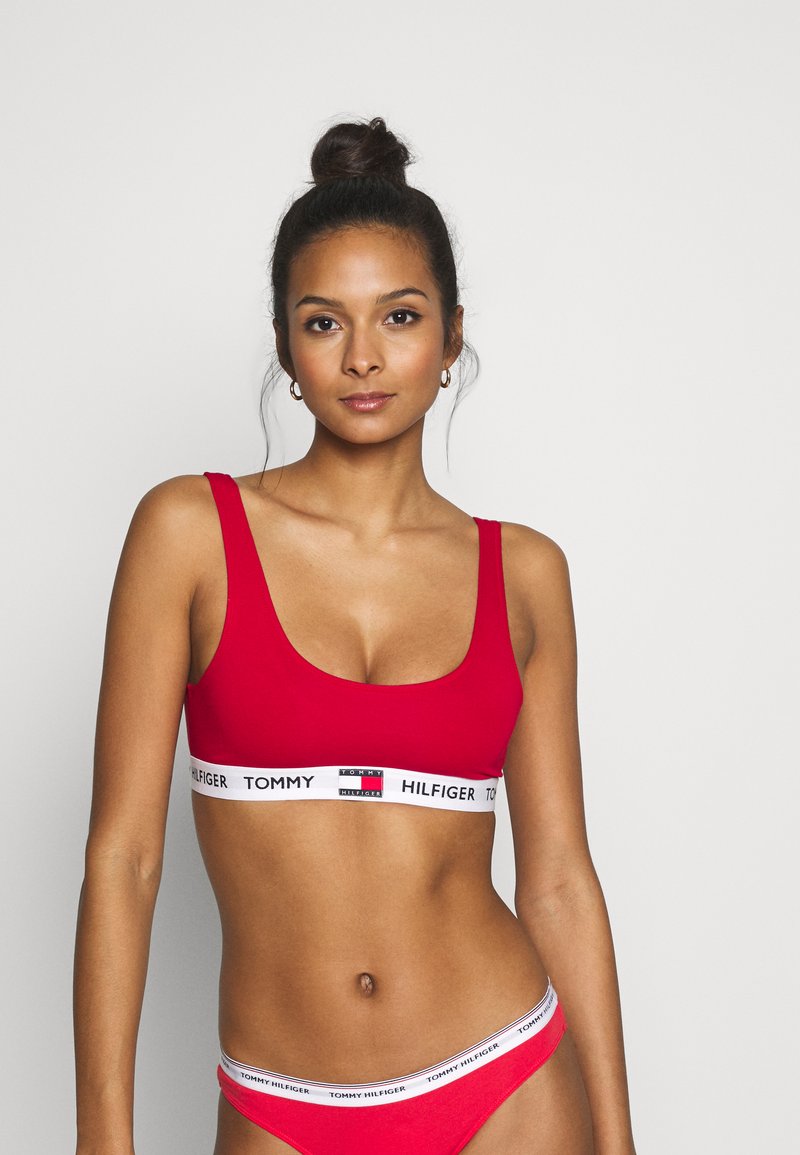 Bralette rossa con una ampia fascia elastica bianca con il testo "TOMMY HILFIGER" in nero. Design semplice con scollatura rotonda e texture liscia. Slip rossi coordinati.