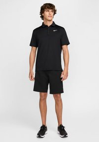 Czarna koszulka polo Nike z kołnierzykiem i zapięciem na trzy guziki, w połączeniu z czarnymi szortami i czarnymi butami sportowymi. Gładka faktura tkaniny.