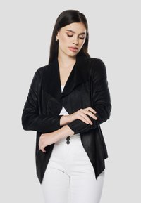 Melnā mākslīgās zamšādas jaka ar platu apkakli, asimetrisku apakšmalu un garām piedurknēm, valkāta pāri baltam topam. Akcentēta ar minimālistiskiem dizaina elementiem.