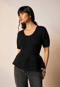 Fekete kötött peplum pulóver rövid ujjakkal, széles kivágással és texturált felülettel. Sötét farmernadrággal és ezüst karkötőkkel párosítva.