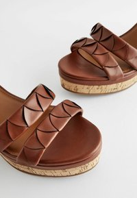 Sandali platform in pelle marrone con doppie cinghie intrecciate dal design ondulato, poggiati su una base in sughero testurizzata. Finitura della superficie liscia.