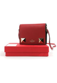 Sac bandoulière Red Valentino Garavani avec des clous pyramides dorés, posé sur une boîte rouge assortie, avec une sangle réglable.