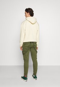 Haut à capuche beige clair avec une grande capuche, associé à un pantalon cargo vert olive comportant plusieurs poches et une coupe ajustée. Des baskets vertes complètent le look.