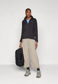 Must zip-up jakiga, kerge kangast, kombineeritud beežide laiade pükstega. Modell hoiab musta seljakotti ja kannab halli matkajalatsit.