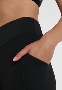 Černé legíny s vysokým pasem, vybavené boční kapsou, vyrobené z elastického materiálu s čistými švy a hladkou texturou.