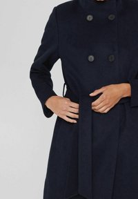 Marineblauwe wollen jas met een dubbelzijdig ontwerp, drie zwarte knopen, een hoge kraag, zijzakken en een stoffen ceintuur.