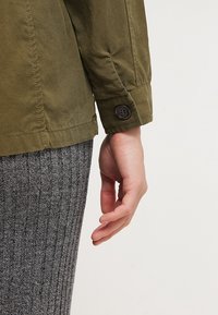 Käsi rentona sivulla, päällä oliivinvihreä takki, jossa napitettu hihansuu ja harmaat ribbineulotut housut yksivärisessä taustassa.