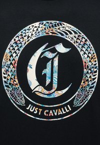 Κυκλικό λογότυπο Just Cavalli με λουλουδάτα σχέδια που σχηματίζουν ένα στυλιζαρισμένο γράμμα "J" και δύο φίδια που περιβάλλουν τον κύκλο σε μαύρο φόντο.