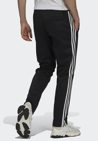 Svarte treningbukser med hvite vertikale striper på sidene. Har elastisk liv og sidelommer. Brukt med lyse sneakers.