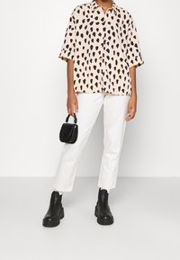 Hellrosa Hemd mit schwarzen unregelmäßigen Punkten, Knopfverschluss, kurze Ärmel. Weiße gerade Hosen und schwarze Leder-Stiefeletten. Kleine schwarze Handtasche.