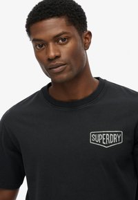 Camiseta de algodón negra con cuello redondo y mangas cortas. En el lado izquierdo del pecho, presenta un logotipo en gris de "SUPERDRY" dentro de una forma de insignia.