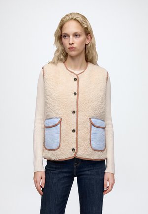 Frau mit blondem Haar trägt eine beige Fleeceweste mit Knöpfen und blauen gesteppte Taschen über einem cremefarbenen Langarmshirt und dunklen Jeans vor weißem Hintergrund.
