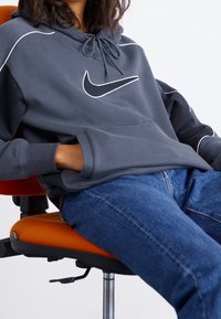 Navy blue hoodie with white stitching and a prominent logo. Features a drawstring hood and kangaroo pocket. Worn with blue jeans.