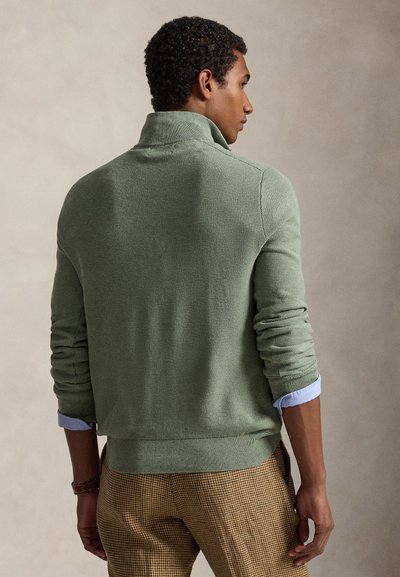 Roheline kootud kampsun kõrge kraega, tekstuurse kangaga, soonikkoes varrukaotste ja äärega, kantud helesinise särgi peal, kombineeritud pruuni ruudulise pükstega.