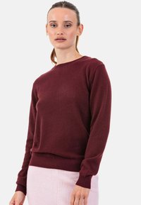 Pull bordeaux à manches longues, col rond et ourlet côtelé. Texture douce, style ajusté, porté sur une jupe rose clair.