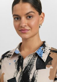 Das mehrfarbige Hemd präsentiert eine Mischung aus beige-, schwarz- und blaufarbigen abstrakten Mustern, mit einem traditionellen Kragen und Knopfdetails. Die glatte Stoffstruktur.