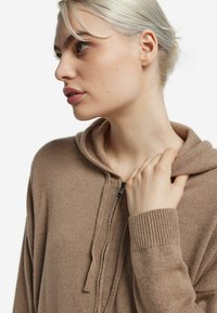 Brązowa bluza z kapturem zapinana na zamek, wykonana z miękkiego materiału, z kapturem i ściągaczami na rękawach. Zdjęcie z bliska podkreślające fakturę i detale kolorystyczne.