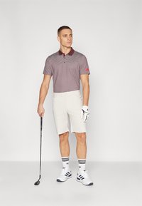 Herrarnas golfklädsel med en kortärmad, murröd och vit mönstrad polo, beiga shorts, vita sportskor och en handske, som håller en golfklubba.