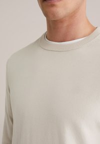 Homme portant un pull beige à col rond par-dessus un sous-maillot blanc, montrant l'encolure et la partie supérieure de la poitrine.