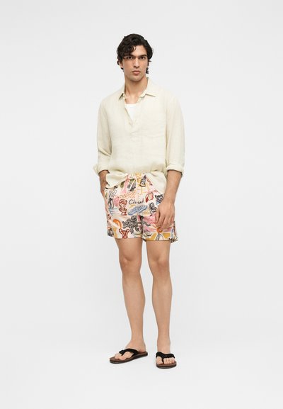 Jeune homme debout portant une chemise beige à manches longues, un short coloré à motifs et des tongs noires sur un fond blanc uni.