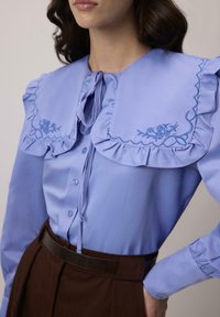 Femeie îmbrăcată cu o bluză bleu deschis, cu un guler mare cu volane brodat cu flori, asortată cu pantaloni maro cu talie înaltă și o curea neagră.