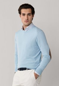 Ung mand iført en lyseblå sweater over en stribet skjorte med krave, hvide bukser og et mørkt bælte, stående med hænderne i lommerne.