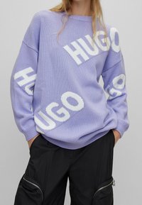 Lavendelkleurige gebreide trui met grote witte "HUGO"-logo's, ronde hals en doorgezakte schouders. Gecombineerd met zwarte cargobroek.