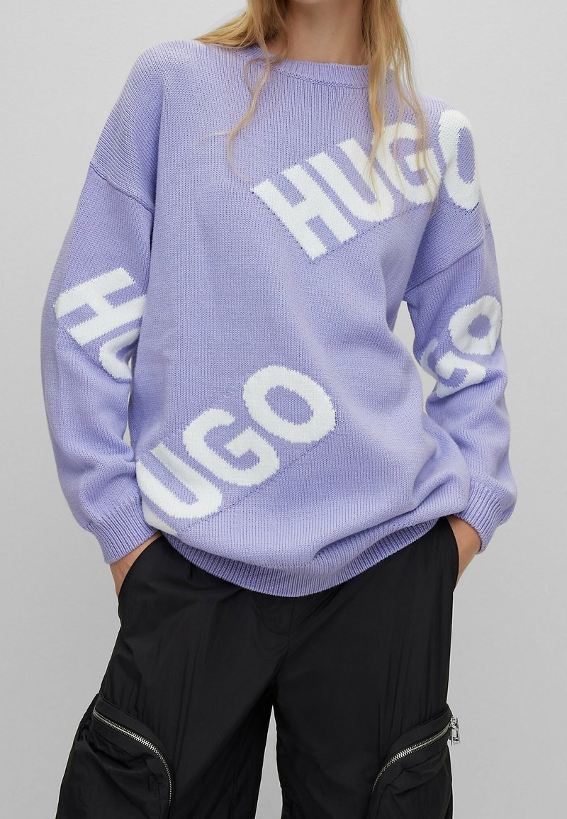Lavendelkleurige gebreide trui met grote witte "HUGO"-logo's, ronde hals en doorgezakte schouders. Gecombineerd met zwarte cargobroek.