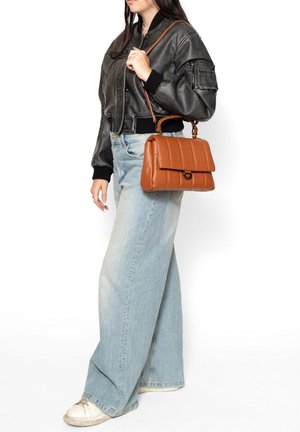 Borsa a tracolla in pelle marrone trapuntata con chiusura a patta e manico superiore, portata su una spalla, abbinata a una giacca bomber nera e jeans blu chiaro.