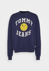 Námorně modrý svetr s kulatým výstřihem, žebrovanými manžetami a lemem. Obsahuje žlutou grafiku usmívající se tváře a text "TOMMY JEANS NY" v bílé barvě.