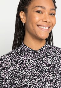 Blusa multicolorida com um padrão abstracto em preto, branco e roxo; fecho frontal com botões e colarinho alto. Tecido com textura suave.