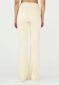 Pantalons beige évasés en denim, dotés d'une taille haute, de poches arrière et d'une texture lisse, avec un léger éclat et des lignes épurées.