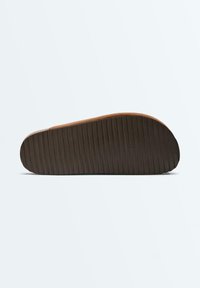 Sandale plate marron avec semelle inférieure sombre texturée et rainurée, vue de dessous sur un fond clair.