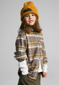 Brązowa koszula w kratę z żółtymi i białymi akcentami, noszona na białej koszuli z długim rękawem. Żółta prążkowana czapka z grafiką gór i logo.