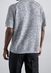 Mężczyzna ubrany w krótki rękaw szary sweter dzianinowy i czarne spodnie, stojący tyłem na jednolitym jasnoszarym tle.