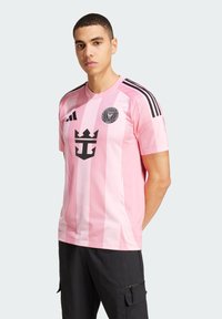 Lyserød kortærmet trøje med sorte striber, der har et krone-logo. Mesh-struktur og rund halsdesign, parret med sorte shorts.