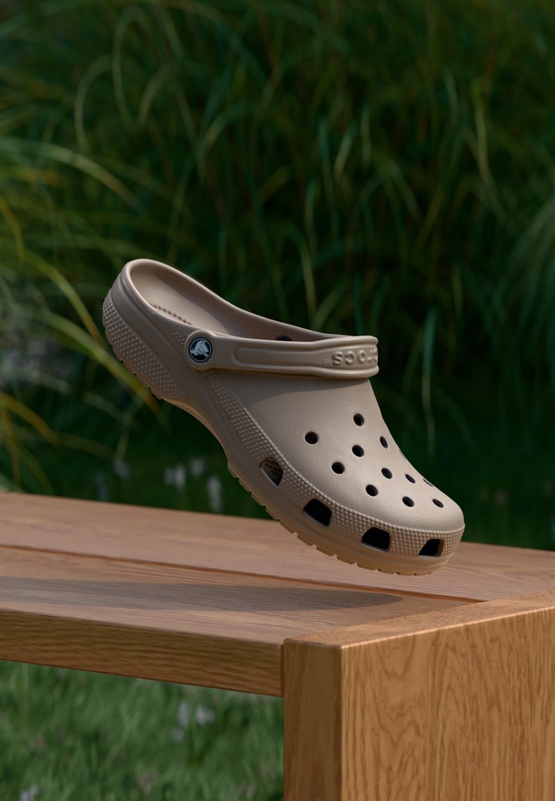 Μπεζ clog στυλ Crocs με οπές αερισμού και λουρί φτέρνας, τοποθετημένο στη γωνία ενός ξύλινου τραπεζιού σε εξωτερικό χώρο, με πράσινα φυτά στο φόντο.