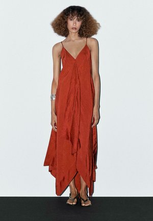 Rusty red, asymmetrical maxi dress with spaghetti straps; features a flowing texture and layered design. Accessorized with a silver cuff bracelet.